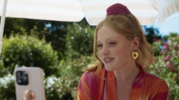 marion-le-corroller-poupée-fondue-téléphone-entretien-2026 Sous un parasol blanc, une femme rousse aux cheveux relevés par un chouchou bordeaux sourit à un téléphone posé devant elle ; elle porte un vêtement satiné aux rayures roses et orangées et de grandes boucles d'oreilles dorées en forme de cœur ; derrière elle, des buissons verdoyants et des fleurs roses sont baignés de soleil ; plan du court-métrage POUPEE FONDUE de Marion Le Corroller.