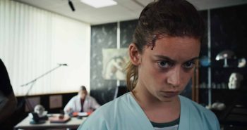marion-le-corroller-mara-taquin-sanguine-entretien-2026 Une jeune femme aux cheveux châtains attachés, vêtue d'une blouse médicale bleu pâle, apparaît en gros plan ; les yeux cernés et le visage fermé, elle fixe intensément quelque chose devant elle ; une plaie sanglante marque sa tempe droite ; à l'arrière-plan, une personne en blouse blanche travaille à un bureau, dans une pièce aux murs sombres où sont exposés des objets anatomiques ; plan du film SANGUINE de Marion Le Corroller.
