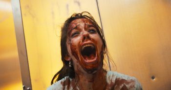 marion-le-corroller-sanguine-kim-higelin-hurlement-entretien-2026 En gros plan, une jeune femme brune au visage ensanglanté renverse la tête en arrière et hurle, la bouche démesurément ouverte ; du sang recouvre son front, ses joues et son cou, tandis que son vêtement blanc est maculé de larges taches rouge sombre ; ses cheveux sont en désordre et ses traits crispés ; derrière elle, une paroi jaune orangé baigne la scène d'une lumière chaude ; plan du film SANGUINE de Marion Le Corroller.