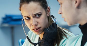 marion-le-corroller-sanguine-pauline-margot-entretien-2026 Une jeune femme en blouse médicale bleu pâle, les cheveux châtains attachés, porte un stéthoscope aux oreilles ; une main gantée de noir maintient l'un des embouts contre son oreille ; les sourcils froncés et les lèvres entrouvertes, elle adresse un regard inquiet à une collègue dont le visage apparaît flou au premier plan, dans une pièce éclairée d'une lumière bleutée ; plan du film SANGUINE de Marion Le Corroller.