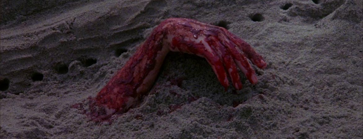 Une main ensanglantée dépasse du sable ; sur une plage ; plan issu du film le silence qui tue de Denny Harris.