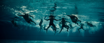 Sous la surface d'une piscine bleu turquoise, une douzaine de garçons en maillot de bain nagent et s'agitent dans toutes les directions ; leurs silhouettes se croisent, les bras et les jambes écartés, au milieu d'une multitude de bulles d'air ; la lumière scintille à la surface, tandis que leurs corps se détachent en ombres sombres sur le bleu profond du bassin ; plan du film THE PLAGUE édité chez Blaq Out.