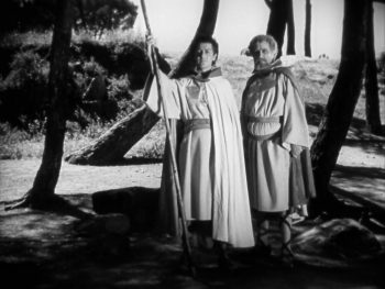Dans un paysage boisé, deux chevaliers en longues tuniques et capes claires regardent au loin ; le premier tient une longue lance dressée à ses côtés ; le second, barbu et légèrement en retrait, porte une épée à la ceinture ; les troncs noirs des arbres contrastent fortement avec l’arrière-plan lumineux ; plan en noir et blanc du film PARSIFAL édité chez Artus Films.