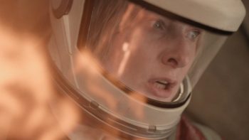 En très gros plan, une femme porte un imposant casque blanc à visière transparente ; les yeux écarquillés et la bouche entrouverte, elle fixe avec inquiétude quelque chose hors champ ; une vive lueur orangée, semblable à des flammes, se reflète sur la visière et envahit le premier plan à gauche ; plan de la saison 3 de la série SILO de Graham Yost.