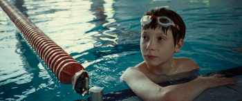 Un jeune garçon aux cheveux bruns mouillés s'accoude au bord d'une piscine, ses lunettes de natation remontées sur le front ; il regarde sur le côté, l'air sérieux, tandis qu'une ligne de séparation rouge et blanche s'étire en diagonale derrière lui, sur l'eau turquoise parcourue de reflets lumineux ; plan du film THE PLAGUE édité chez Blaq Out.