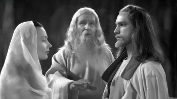 Une jeune femme voilée comme la Vierge Marie et un jeune homme aux longs cheveux se font face ; entre eux, un vieillard à la longue barbe blanche tend les mains dans leur direction ; les yeux baissés, la femme reste immobile tandis que le jeune homme la regarde, leurs silhouettes claires se détachant sur un fond sombre ; plan en noir et blanc du film PARSIFAL édité chez Artus Films.