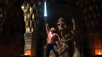 Dans une salle monumentale aux murs de pierre sombre, un jeune homme blond en chemise rose brandit une immense épée lumineuse au-dessus de sa tête ; les jambes écartées et le visage tourné vers la lame, il se tient devant une gigantesque sculpture de crâne aux orbites creuses, encadrée de colonnes massives ; deux braseros éclairent la salle de leurs flammes orangées, tandis que l'épée projette une vive lumière blanche ; plan du film LES MAITRES DE L'UNIVERS de Travis Knight.