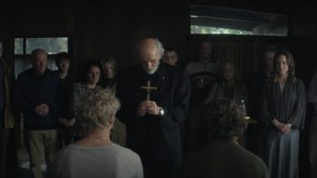 Dans une pièce sombre , un prêtre à la barbe blanche habillé tout en noir incline la tête en avant vers les deux adolescents face à lui ; une croix tenu dans ses deux mains ; avec des fidèles autour de lui ; dans une pièce sombre ; plan issu du film Leviticus de Adrian Chiarella.