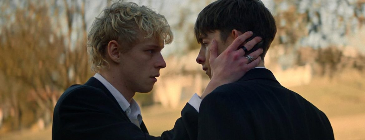 Deux adolescents face à face, un blond caresse de sa main droite la joue du brun ; avec en arrière plan une forêt ; plan issu du film Leviticus de Adrian Chiarella.