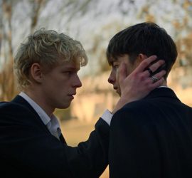 Deux adolescents face à face, un blond caresse de sa main droite la joue du brun ; avec en arrière plan une forêt ; plan issu du film Leviticus de Adrian Chiarella.