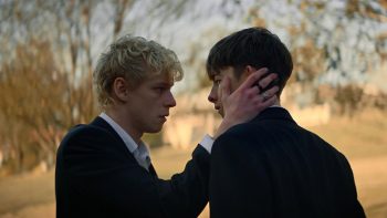 Deux adolescents face à face, un blond caresse de sa main droite la joue du brun ; avec en arrière plan une forêt ; plan issu du film Leviticus de Adrian Chiarella.
