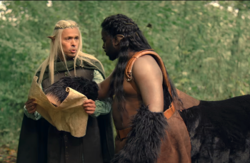 Éric Judor, vêtu d'une cape verte et portant de longs cheveux blonds et des oreilles pointues, consulte un parchemin aux côtés d'un centaure dans Le Roi du Game.