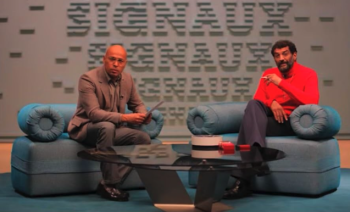 Éric Judor en costume gris et Ramzy Bedia en pull rouge assis sur des canapés bleus face à une table basse, dans Signaux de Quentin Dupieux.