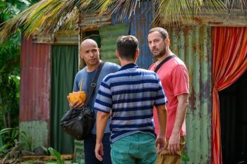Éric Judor et Fred Testot discutent avec un troisième homme devant des habitations colorées entourées de végétation tropicale dans la série Platane.