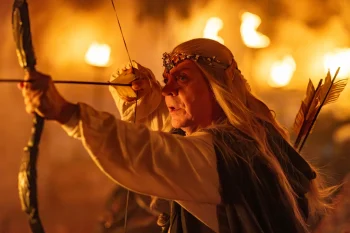 Marc Labrèche en costume d'elfe aux longs cheveux blonds, visant avec un arc dans une scène enflammée du film Le Roi du Game d'Éric Judor.