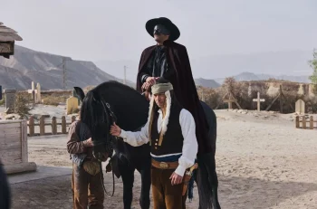 Un cavalier masqué vêtu d'une cape sombre est assis sur un cheval noir, accompagné d'un homme en costume de western dans un décor de cimetière désertique, dans Tout simplement fan.